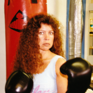 1980s era photo of Suzanne Curran with long curly brown hair and black boxing gloves standing in front of a red century punching bag.